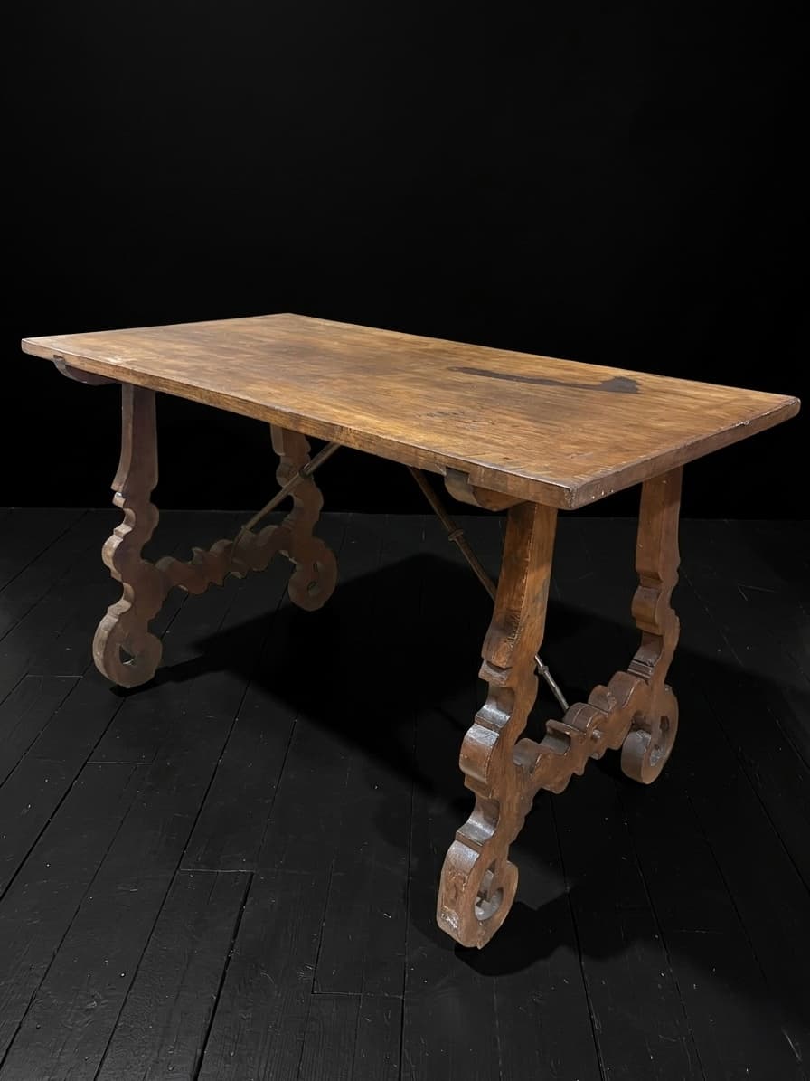 Mesa rústica de madera con patas talladas de estilo lira y tirantes de hierro.